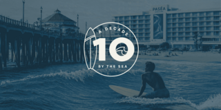 A surfer rides a wave near a beach, with a bustling pier and resort in the background under a bright sky. Palm trees line the shore.