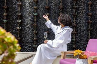 A woman in a white robe sits peacefully, holding a glass of champagne, near a textured wall adorned with hanging features, enjoying calm moments.