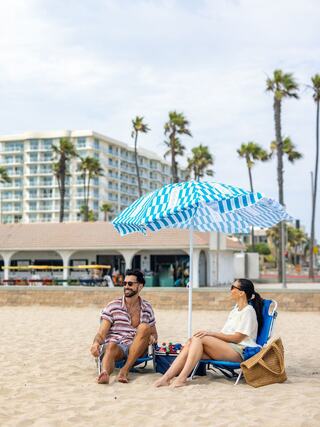 A couple relaxes on the beach under a striped umbrella, enjoying the sun with palm trees and a hotel in the background.