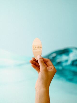 A hand holds a wooden keycard with "Pasea Hotel & Spa" engraved, set against a soft blue ocean-themed background.