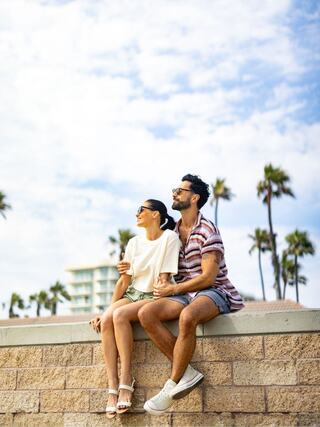 A couple sits on a wall, enjoying each other's company, with palm trees and a sunny sky in the background, radiating happiness and relaxation.