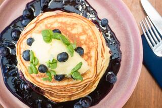 Stacked pancakes are topped with blueberries and a dollop of cream, all drizzled with rich blueberry syrup, garnished with fresh basil.