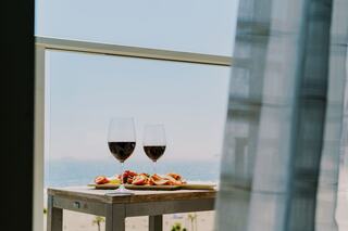 Two glasses of red wine sit on a wooden table alongside a platter of snacks, with a serene coastal view in the background.