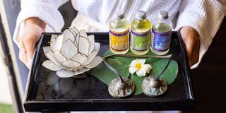 A black tray holds a decorative lotus, three small bottles of oils, a green leaf, and a white flower, creating a serene, spa-like presentation.