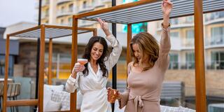 Two women celebrate outdoors, enjoying drinks and having fun under stylish wooden structures, with a modern backdrop in a lively atmosphere.