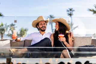 A cheerful couple in cowboy hats relaxes on a couch, enjoying colorful cocktails with palm trees and sunshine in the background.