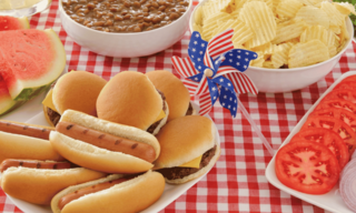 A festive spread features mini hamburgers and hot dogs, alongside baked beans, chips, watermelon, and a decorated pinwheel on a checkered tablecloth.