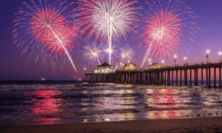 Colorful fireworks burst over a pier, reflecting in the water as the sky transitions to twilight, creating a festive seaside atmosphere.