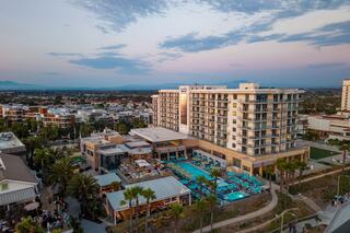 A modern hotel with a large pool area, surrounded by palm trees and cityscape, under a pastel sky at dusk.
