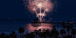 Colorful fireworks illuminate the sky above a pier, reflecting on the ocean waves as twilight sets in, creating a festive atmosphere.