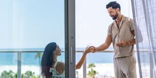 A man in casual attire offers a drink to a woman sitting on a balcony, with a stunning ocean view in the background.