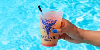 A hand holds a vibrant cocktail in a branded cup, contrasting against a sparkling blue swimming pool backdrop.