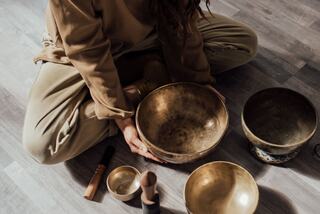 A person sits cross-legged, surrounded by various bowls and instruments, engaged in a soothing practice or meditation.