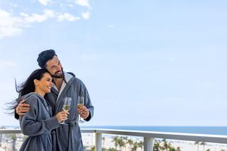 A happy couple in cozy robes toasts with champagne glasses on a balcony, overlooking a scenic coastal view.