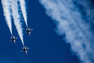 A crowd faces the sky, watching smoke trails above palm trees on a sunny day, creating a festive atmosphere at a seaside location.