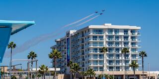 A formation of jet planes creates smoke trails above a beachfront hotel, framed by palm trees under a clear blue sky.