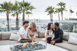Four friends enjoy drinks and laughter around a cozy fire pit, surrounded by palm trees and a beachfront view.