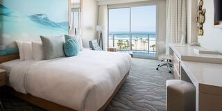 A modern hotel room features a large bed, coastal artwork, and a breathtaking ocean view through expansive glass doors.