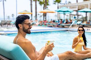 A man enjoys a cocktail by the pool, smiling at a woman in a yellow swimsuit, surrounded by sun loungers and palm trees.