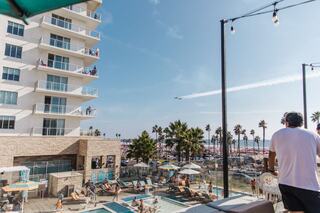 A sunny coastal scene featuring a hotel, a pool area with loungers, palm trees, and an ocean view, with people enjoying the atmosphere.