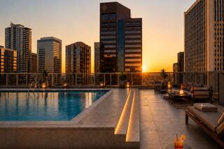 A rooftop pool glows at sunset, surrounded by modern skyscrapers. Cozy seating and drinks create a relaxing urban oasis.