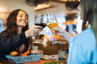 Two women toast with wine glasses, smiling joyfully over a table filled with delicious food in a vibrant restaurant setting.