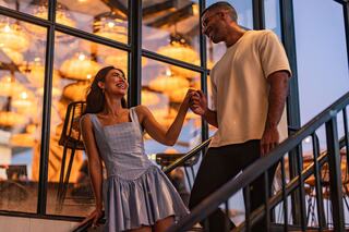 A couple shares a joyful moment, holding hands and smiling, surrounded by warm, glowing lights in a stylish setting.