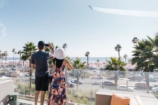 A couple stands on a balcony, admiring a performance in the sky above a sunny beach lined with palm trees and parked cars.