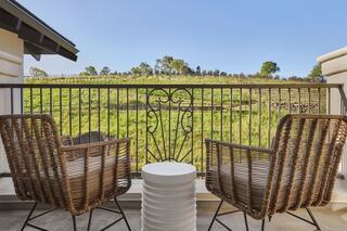 A cozy balcony features two wicker chairs and a small table, overlooking lush green fields and hills under a clear blue sky.