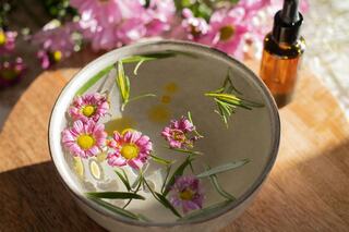 A bowl filled with water showcasing pink flowers and green leaves, accompanied by a small amber bottle, evokes a serene, natural vibe.