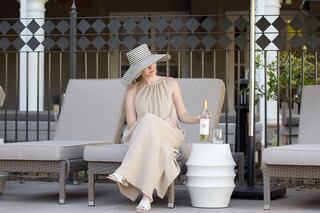 A woman in a flowing beige dress and wide-brimmed hat relaxes on a pool lounge chair, sipping wine beside a stylish white table.