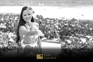 A woman in a floral dress, adorned with jewelry and a flower in her hair, smiles and gestures warmly against a scenic backdrop.