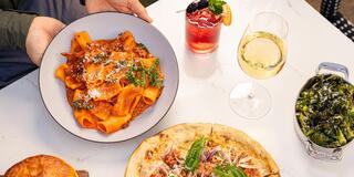 A delicious spread featuring pasta in a rich sauce, a refreshing cocktail, white wine, pizza topped with fresh ingredients, and sautéed greens.