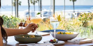A coastal dining scene with colorful cocktails and a vibrant salad, framed by palm trees and a sunny beach view. Relaxing and inviting atmosphere.
