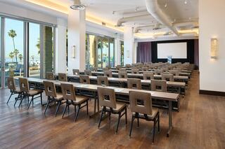 A spacious conference room features rows of tables and chairs, large windows with palm trees, and a screen at the front for presentations.