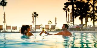 A couple joyfully clinks glasses in a pool at sunset, surrounded by palm trees and lounge chairs, creating a relaxed, romantic atmosphere.