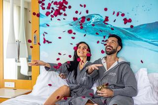 A joyful couple in robes tosses rose petals while enjoying drinks in a stylish hotel room, surrounded by a serene ocean mural.