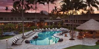 A serene resort pool area surrounded by palm trees and lounge chairs, set against a beautiful sunset, evokes relaxation and tranquility.