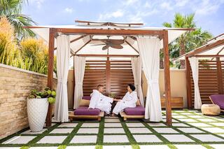 Two individuals relax in cozy robes under a shaded cabana, enjoying drinks amidst lush greenery and a serene atmosphere.