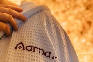 A hand holds a textured white robe featuring the logo "arna spa," with a warm, softly lit background suggesting a relaxing atmosphere.