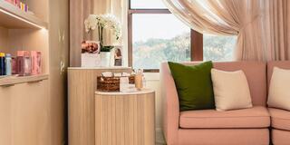 A cozy, elegant seating area features a pink sofa with cushions, a round side table, and a window draped in soft fabric, adding warmth to the space.