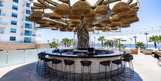 A stylish outdoor bar features a statue of Liberty under woven canopies, surrounded by palm trees and a scenic coastal view.