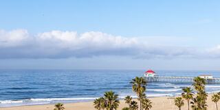 A serene beach scene with gentle waves, palm trees, and a pier featuring a red-roofed structure under a clear blue sky.