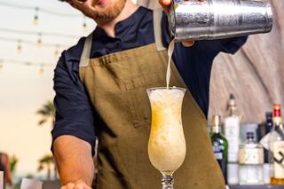 A bartender in an apron pours a cocktail from a shaker into a glass, surrounded by bottles and outdoor string lights.