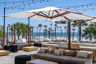 A stylish outdoor lounge features comfy seating, palm trees, and ocean views, enhanced by string lights and a large umbrella.