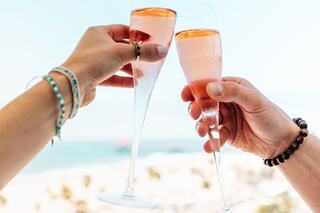 Two hands clink elegant flutes filled with sparkling pink drinks, set against a sunny beach backdrop. Celebratory vibes abound!