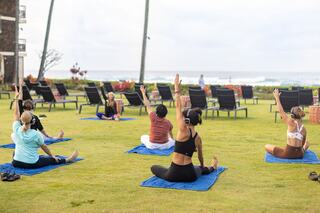A group of individuals practices yoga outside on a green lawn, with ocean views and headphones, engaging in a serene workout.