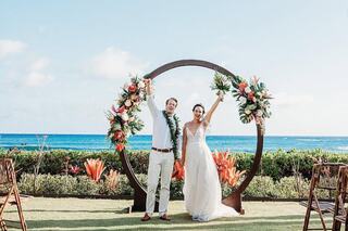 A joyful couple celebrates their wedding under a floral arch, with the ocean and a clear sky in the background, surrounded by nature.