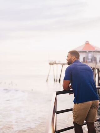 A man in a blue shirt leans on a railing, gazing out at the ocean and pier during a serene, cloudy sunset.
