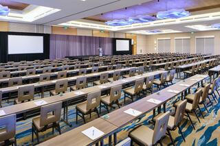 A spacious conference room with rows of tables and chairs, equipped for a business or seminar event, featuring presentation screens at the front.